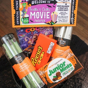 May include: A black bucket filled with Halloween-themed treats, including a movie ticket, a box of Reese's Pieces, a bag of Junior Mints, and a bag of green pretzel rods. The ticket reads "Welcome to Thrills & Chills Movie Theater".