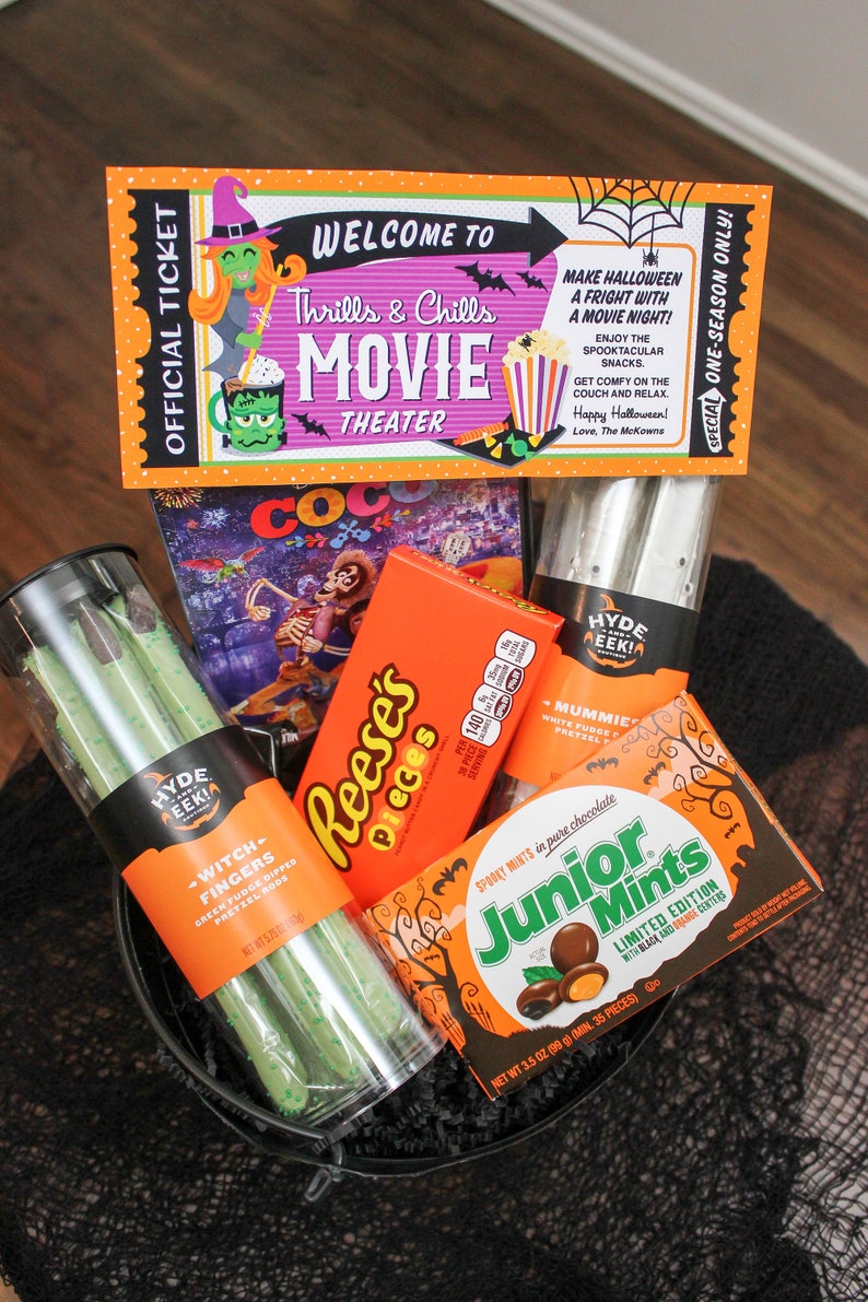 May include: A black bucket filled with Halloween-themed treats, including a movie ticket, a box of Reese's Pieces, a bag of Junior Mints, and a bag of green pretzel rods. The ticket reads "Welcome to Thrills & Chills Movie Theater".