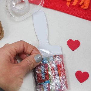 May include: A clear plastic bag filled with colorful candy, held near a white paint palette-shaped card. Two red heart-shaped cutouts are nearby. A clear tape dispenser and red fringed paper are in the background.