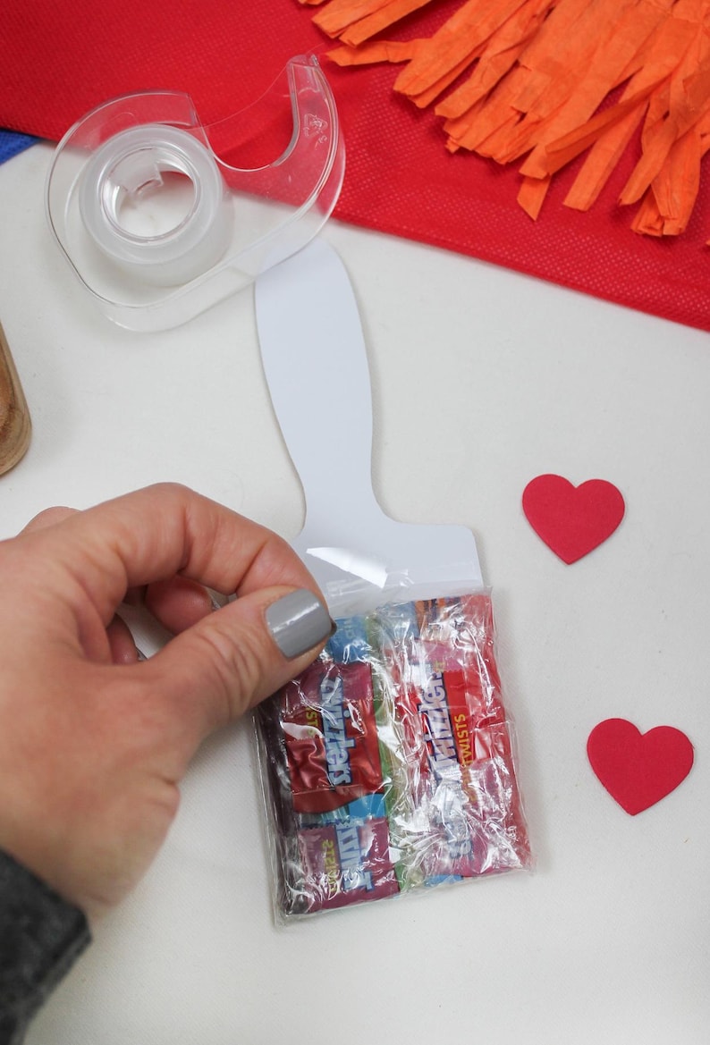 May include: A clear plastic bag filled with colorful candy, held near a white paint palette-shaped card. Two red heart-shaped cutouts are nearby. A clear tape dispenser and red fringed paper are in the background.