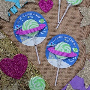 Op de afbeelding: Drie groene en witte swirl-lollipops met ruimtethema-verpakkingen. De verpakkingen hebben een roze planeetring, een ruimteschip en de tekst "Valentine. You're out of this world!" en "I'm so happy we share the same space!" met de naam "Sawyer" onderaan.
