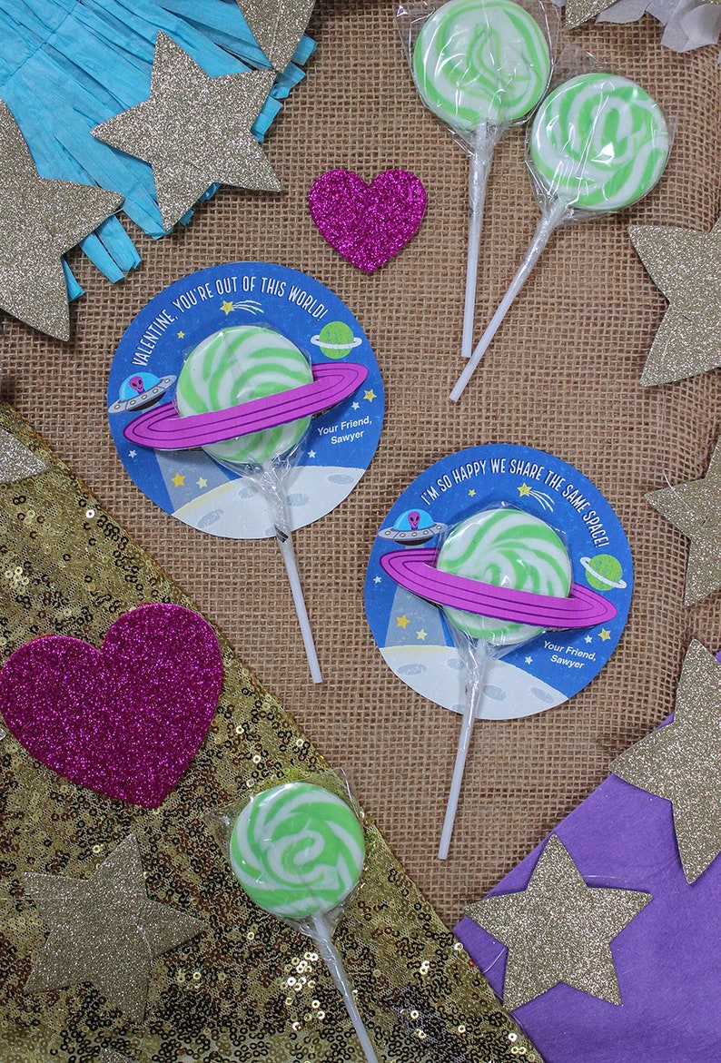 Op de afbeelding: Drie groene en witte swirl-lollipops met ruimtethema-verpakkingen. De verpakkingen hebben een roze planeetring, een ruimteschip en de tekst "Valentine. You're out of this world!" en "I'm so happy we share the same space!" met de naam "Sawyer" onderaan.