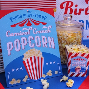 May include: A blue and red sign with the text "Proud Purveyor of Carnival Crunch Popcorn" with a red and white striped popcorn container and stars. A glass jar filled with popcorn and a red and white striped popcorn container with the text "Carnival Crunch Popcorn" are on a blue and red table.