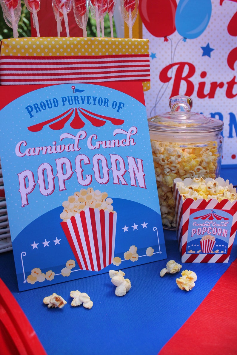 May include: A blue and red sign with the text "Proud Purveyor of Carnival Crunch Popcorn" with a red and white striped popcorn container and stars. A glass jar filled with popcorn and a red and white striped popcorn container with the text "Carnival Crunch Popcorn" are on a blue and red table.
