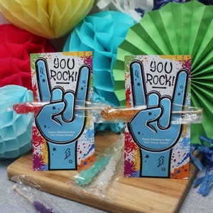 Pode incluir: Dois cart&otilde;es coloridos de Dia dos Namorados com o texto "You Rock!" e um gesto de m&atilde;o de rock and roll. Cada cart&atilde;o tem um palito de pirulito de rocha preso a ele. Os cart&otilde;es est&atilde;o sobre uma superf&iacute;cie de madeira com papel de seda colorido no fundo.