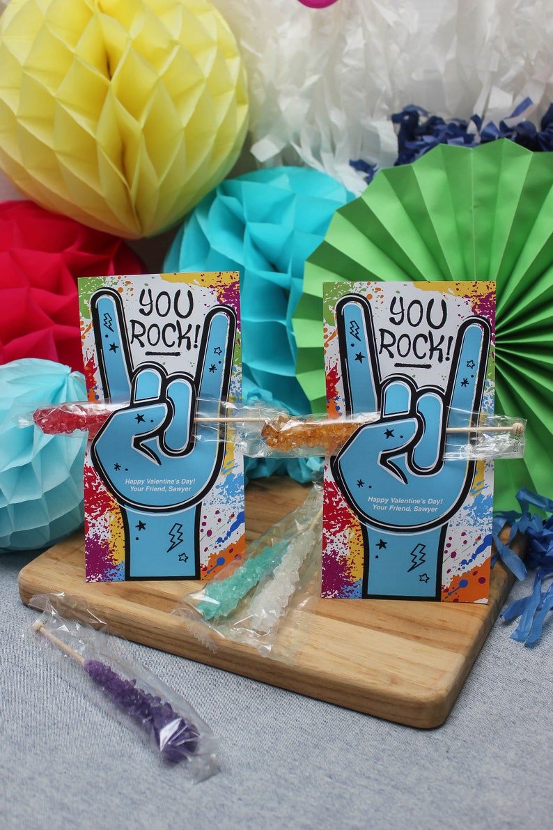 Pode incluir: Dois cart&otilde;es coloridos de Dia dos Namorados com o texto "You Rock!" e um gesto de m&atilde;o de rock and roll. Cada cart&atilde;o tem um palito de pirulito de rocha preso a ele. Os cart&otilde;es est&atilde;o sobre uma superf&iacute;cie de madeira com papel de seda colorido no fundo.