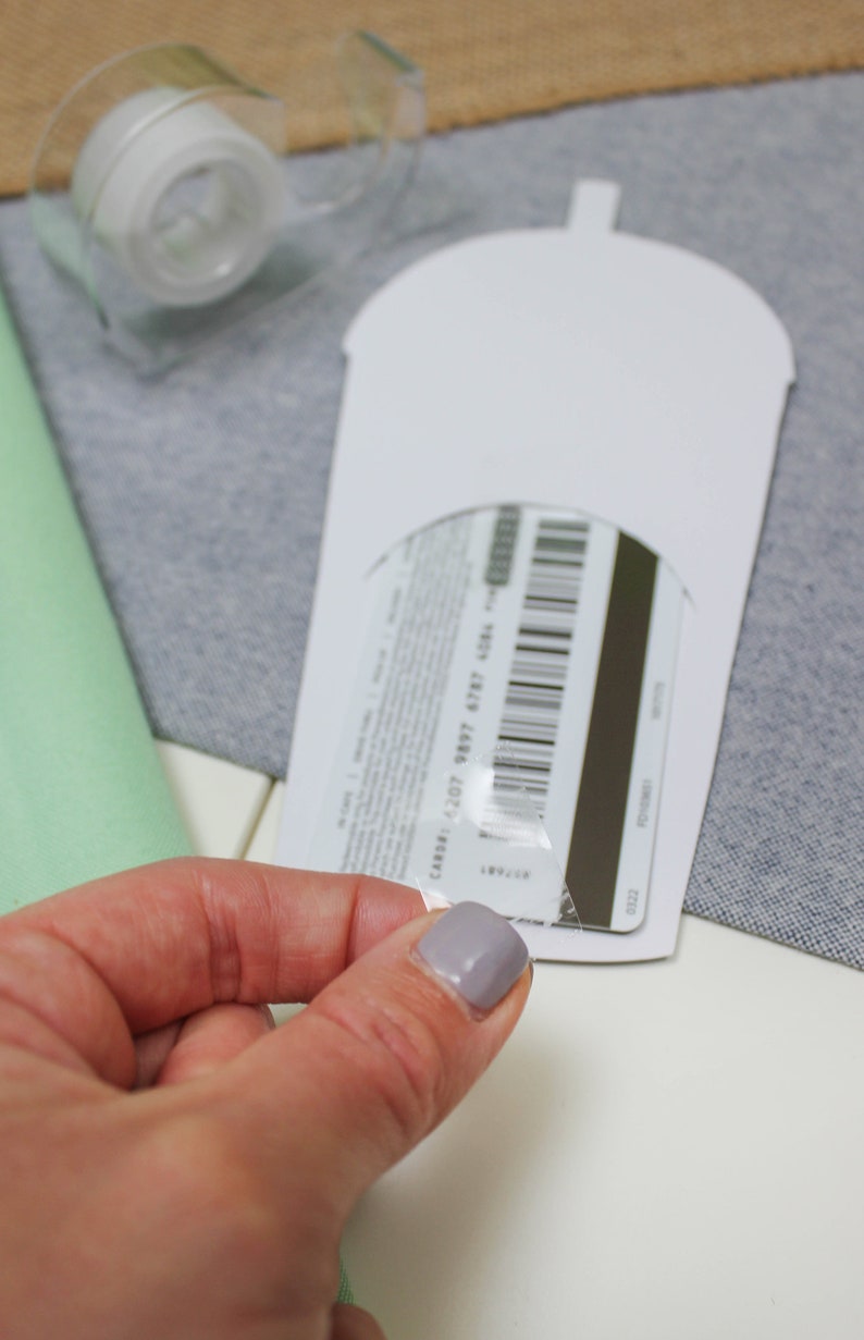 May include: A white cardboard cutout of a cup with a clear plastic gift card inside. The gift card has a barcode and a black stripe. The gift card is being peeled off of the plastic.