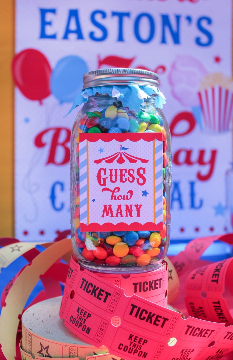 May include: A glass jar filled with colorful candy, with a red, white, and blue circus-themed label that says "Guess how many". The jar is sitting on top of a stack of red paper tickets that say "Ticket" and "Keep this coupon".
