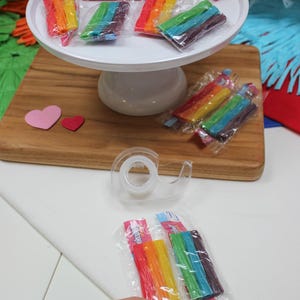 May include: Rainbow-colored candy sticks in clear plastic packaging are displayed on a white cake stand and a wooden cutting board. A red bag with text is visible. The text on the bag reads: BIG BAGS. BIGGER FLAVORS. ZERO JUDGMENT.