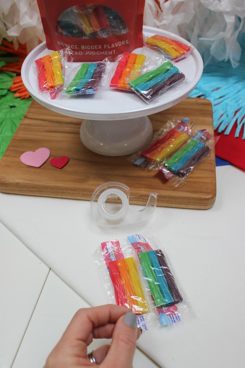 May include: Rainbow-colored candy sticks in clear plastic packaging are displayed on a white cake stand and a wooden cutting board. A red bag with text is visible. The text on the bag reads: BIG BAGS. BIGGER FLAVORS. ZERO JUDGMENT.