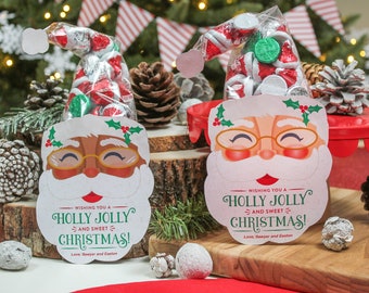 May include: Two Santa Claus-themed gift bags filled with candy. The bags are made of clear plastic and have a white Santa Claus face with a red nose and black eyes. The bags are decorated with holly and a green bow. The text on the bags reads "Wishing you a Holly Jolly and Sweet Christmas! Love, Sawyer and Easton."