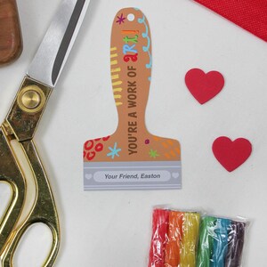 May include: A craft-themed image featuring a paint brush-shaped card with the text "YOU'RE A WORK OF ART!" and "Your Friend, Easton". Two red heart cutouts, a pair of gold scissors, and a package of rainbow-colored craft sticks are also visible.