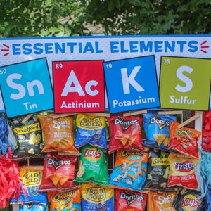 Puede incluir: Un cartel con las palabras "Essential Elements" e imágenes de elementos químicos con los nombres "Sn", "Ac", "K" y "S" que representan "Tin", "Actinium", "Potassium" y "Sulfur".  Se muestran una variedad de bolsas de aperitivos debajo del cartel.