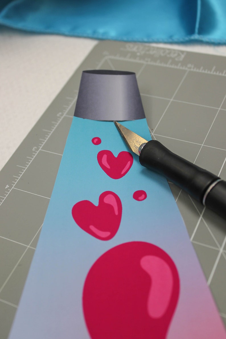 Peut inclure: Un projet d'artisanat de lampe &agrave; lave bleue et rose avec un motif en forme de c&oelig;ur. Un couteau artisanal est utilis&eacute; pour couper le papier.