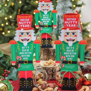 May include: Three glass jars filled with mixed nuts are decorated with nutcracker-themed gift tags. The tags feature a cartoon nutcracker with the text "Another Nutty Year" and a holiday message. The jars are sitting on a red surface.