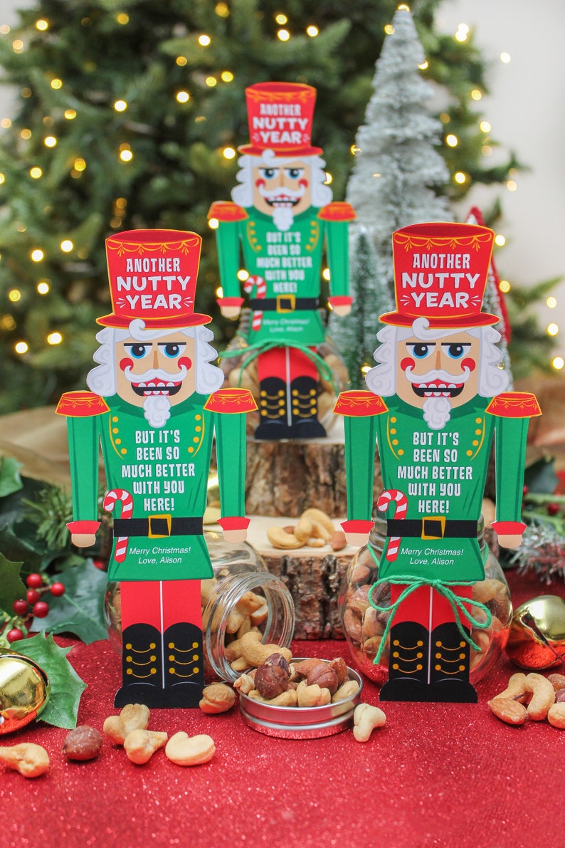 May include: Three glass jars filled with mixed nuts are decorated with nutcracker-themed gift tags. The tags feature a cartoon nutcracker with the text "Another Nutty Year" and a holiday message. The jars are sitting on a red surface.