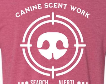May include: A maroon t-shirt with a white graphic design. The design features a dog nose inside a target, with the words "CANINE SCENT WORK" above and "SEARCH ALERT!" below. A star and dashes complete the design.