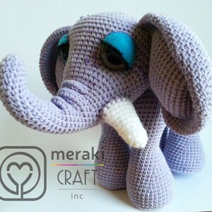 Könnte beinhalten: Ein gehäkeltes lilafarbenes Elefantenspielzeug mit weißen Stoßzähnen und blauen Augen. Der Elefant steht auf einer weißen Oberfläche. Das Spielzeug ist handgefertigt und hat einen verspielten Look.