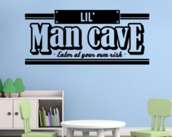 May include: A black and white vinyl wall decal with the text "Lil' Man Cave - Enter at your own risk -" on a light blue wall. The decal is above a white table and two green chairs in a room with a white bookshelf.