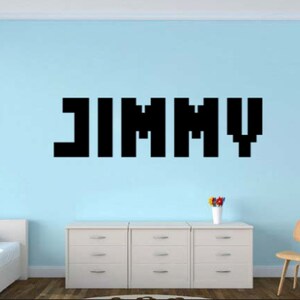 May include: A light blue wall with a black pixelated name "JIMMY" on it. There is a white dresser with three drawers and a chair in the room.