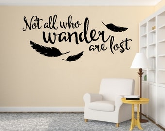Puede incluir: Una calcomanía de pared con la cita "Not all who wander are lost" en escritura negra con siluetas de plumas negras. La calcomanía está en una pared beige con un sillón blanco y una pequeña mesa auxiliar amarilla.