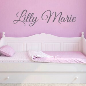 May include: A white daybed with a pink and white patterned bedspread and a pink pillow. The wall behind the bed is light pink with the name "Lilly Marie" written in gray cursive lettering.