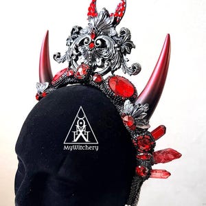 May include: A gothic-style crown with red and silver accents. The crown features large red horns, red gemstones, and intricate silver filigree. The text "MyWitchery" is printed on the front.