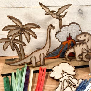 May include: A wooden craft kit featuring a dinosaur, a palm tree, a volcano, and a pterodactyl. The kit includes colorful glass tiles and a paintbrush for creating a mosaic art project.
