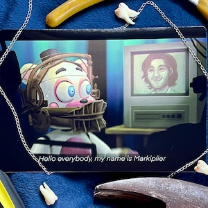 May include: A white animatronic character with a pink face and a metal jaw trap. The character is looking at a computer screen with a man's face on it. The text on the screen says "Hello everybody, my name is Markiplier".