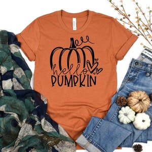 Puede incluir: Una camiseta naranja quemada con un gráfico negro de una calabaza y el texto "hello pumpkin" con corazones.