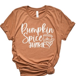 Op de afbeelding: Een bruin T-shirt met de tekst "Pumpkin Spice Junkie" in witte letters. Naast de tekst staat een witte omtrek van een koffiebeker met een pompoen en een hart.