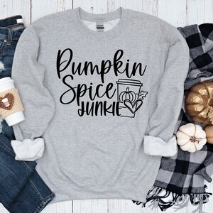 Peut inclure: Un sweat-shirt gris avec le texte "Pumpkin Spice Junkie" en lettres noires. Le sweat-shirt est posé sur une surface en bois blanc avec un jean bleu, une tasse à café et une couverture à carreaux avec des citrouilles dorées.