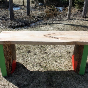 Peut inclure: Un banc en bois avec un plateau en bois naturel et des pieds peints en vert et rouge. Le banc est posé sur une surface herbeuse.