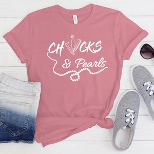 May include: A pink t-shirt with the text "Chucks & Pearls" in white lettering. The text is surrounded by a white pearl necklace graphic.