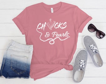 May include: A pink t-shirt with the text "Chucks & Pearls" in white lettering. The text is surrounded by a white pearl necklace graphic.