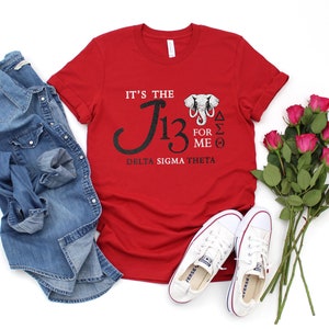 May include: A red t-shirt with the text "IT'S THE J13 FOR ME" and "DELTA SIGMA THETA" printed on it. The shirt is paired with a denim shirt, white sneakers, and a bouquet of pink roses. The shirt has a graphic of an elephant.