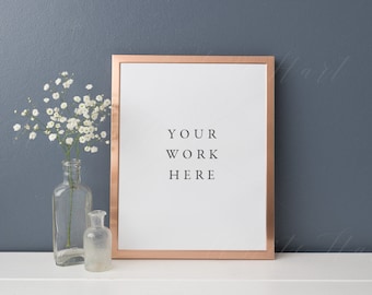 May include: A copper-colored frame with a white mat and a white background. The text "YOUR WORK HERE" is printed in black on the white background. A small bouquet of white baby's breath flowers is in a clear glass vase to the left of the frame. The frame is sitting on a white surface.