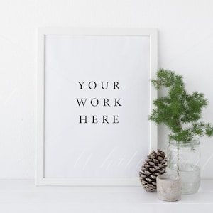 May include: White frame with a white mat and the text "YOUR WORK HERE" inside. A small pine cone and a sprig of greenery in a glass jar are on a white surface in front of the frame.