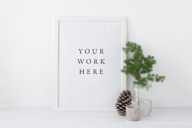 May include: White frame with a white mat and the text "YOUR WORK HERE" inside. A small pine cone and a sprig of greenery in a glass jar are on a white surface in front of the frame.