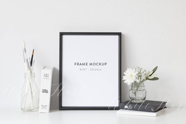 May include: A black frame mockup with a white background. The frame is 8x10 inches or 20x25 centimeters. The frame is sitting on a white surface with a book, a vase of white flowers, and a glass jar with paintbrushes.
