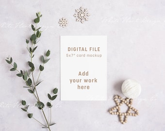 May include: A white card mockup with the text "DIGITAL FILE 5x7" card mockup Add your work here". The card is surrounded by a sprig of greenery, a white wooden ball, a wooden star, and two small white wooden snowflakes.