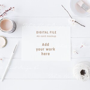 May include: A white card mockup with the text "DIGITAL FILE A6 card mockup Add your work here" on a white wooden background with paint jars, a paintbrush, and white flowers.