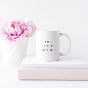 Op de afbeelding: Een witte keramische mok met een zwarte inscriptie die "ADD YOUR DESIGN" luidt op een witte achtergrond. De mok staat op een stapel boeken met roze pioenen in een witte vaas.