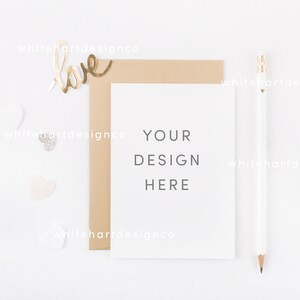 Puede incluir: Una tarjeta blanca con el texto "YOUR DESIGN HERE" sobre un fondo blanco con un sobre marrón, un lápiz dorado y corazones de purpurina dorada.