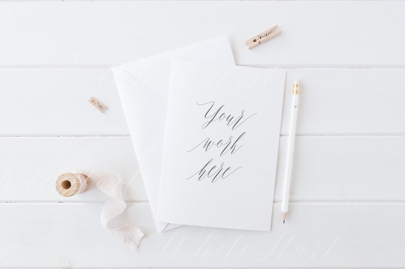 K&ouml;nnte beinhalten: Eine wei&szlig;e Karte mit dem Text "Your work here" in schwarzer Kalligraphie. Die Karte liegt auf einer wei&szlig;en Holzoberfl&auml;che mit einem wei&szlig;en Umschlag, einem Bleistift und einer Spule wei&szlig;en Bandes.
