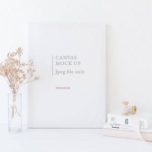 May include: White canvas mock-up with the text "Canvas Mock Up Jpeg file only 30x40CM" on a white background. A vase with dried flowers and a spool of thread are on a white surface in front of the canvas.