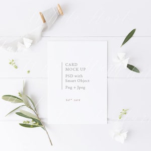 May include: A white 5x7" card mockup with text, surrounded by decorative elements. The card features the text "CARD MOCK UP PSD with Smart Object Png + Jpeg". A white ribbon, sprigs, and olive branches are arranged on a white wooden surface.