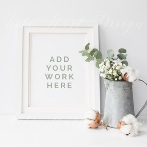 May include: White picture frame with a white mat and the text "ADD YOUR WORK HERE" inside. The frame is sitting on a white surface with a metal pitcher filled with white flowers and cotton bolls.