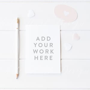 Puede incluir: Una tarjeta blanca con el texto "ADD YOUR WORK HERE" sobre un fondo de madera blanca con un lápiz y corazones rosas.