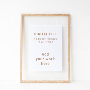 Pu&ograve; includere: Una cornice A3 in legno chiaro con un mockup di carta A4 bianca. Il testo sulla carta recita "DIGITAL FILE, A4 paper mockup in A3 frame, Add your work here."